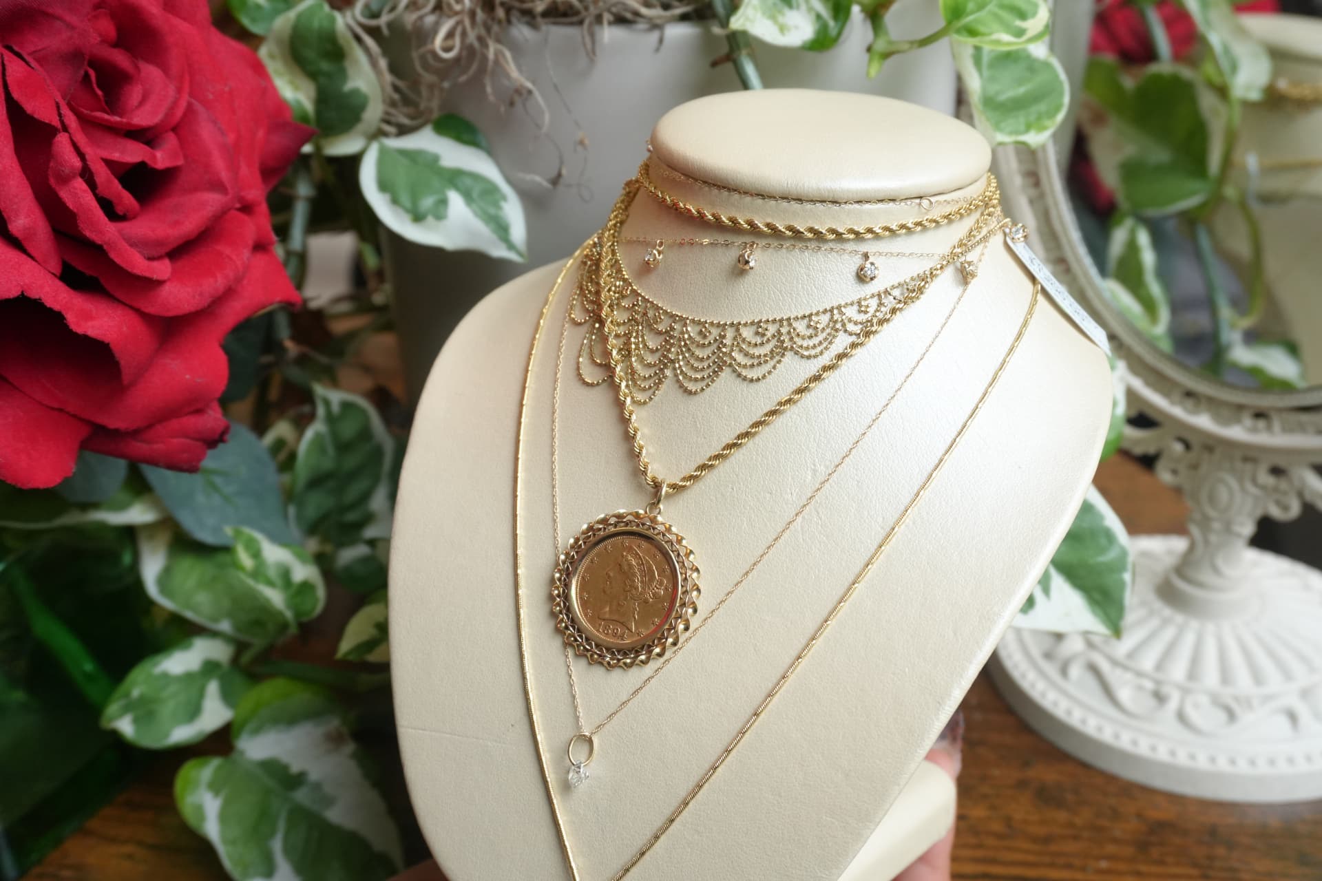 Layered gold chains with a framed gold coin pendant on a cream bust beside a red rose